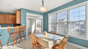 A dining area with a table set for four, wooden chairs, a kitchen with wooden cabinets, and a large window with blinds. - 🏖️AMAZING 4 Bd, West Ocean City 🏖️ (Ocean City)
