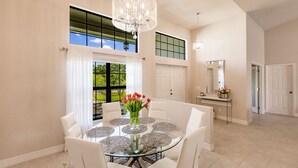 Un vaste espace dédié à la salle à manger, avec une table en verre entourée de chaises blanches, un lustre et une vue sur la verdure à l’extérieur.