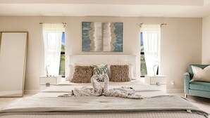 Une chambre avec un grand lit, deux tables de chevet, un fauteuil turquoise et un tableau au-dessus du lit.