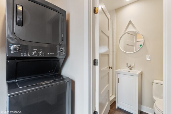 A compact laundry room with a front-loading washer and dryer, a white door leading to a bathroom with a sink and toilet.