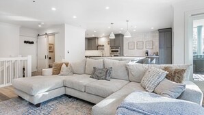 Una sala moderna con un amplio sofá seccional en forma de L, una cocina con armarios grises e iluminación colgante.