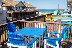 A deck with blue chairs and a table, overlooking a beach and a building with balconies.
