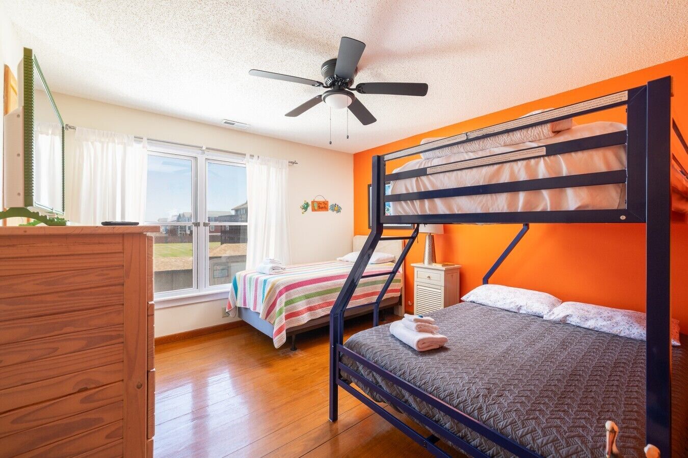 A bunk bed room with a ceiling fan, a window with curtains, a wooden dresser, and a view of the outside.