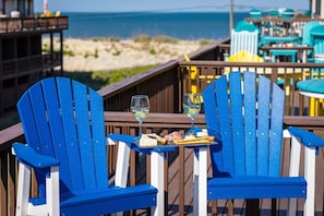 Dos sillas Adirondack azules con una mesa entre ellas, una copa de vino blanco, y una tabla de quesos con galletas y embutidos.