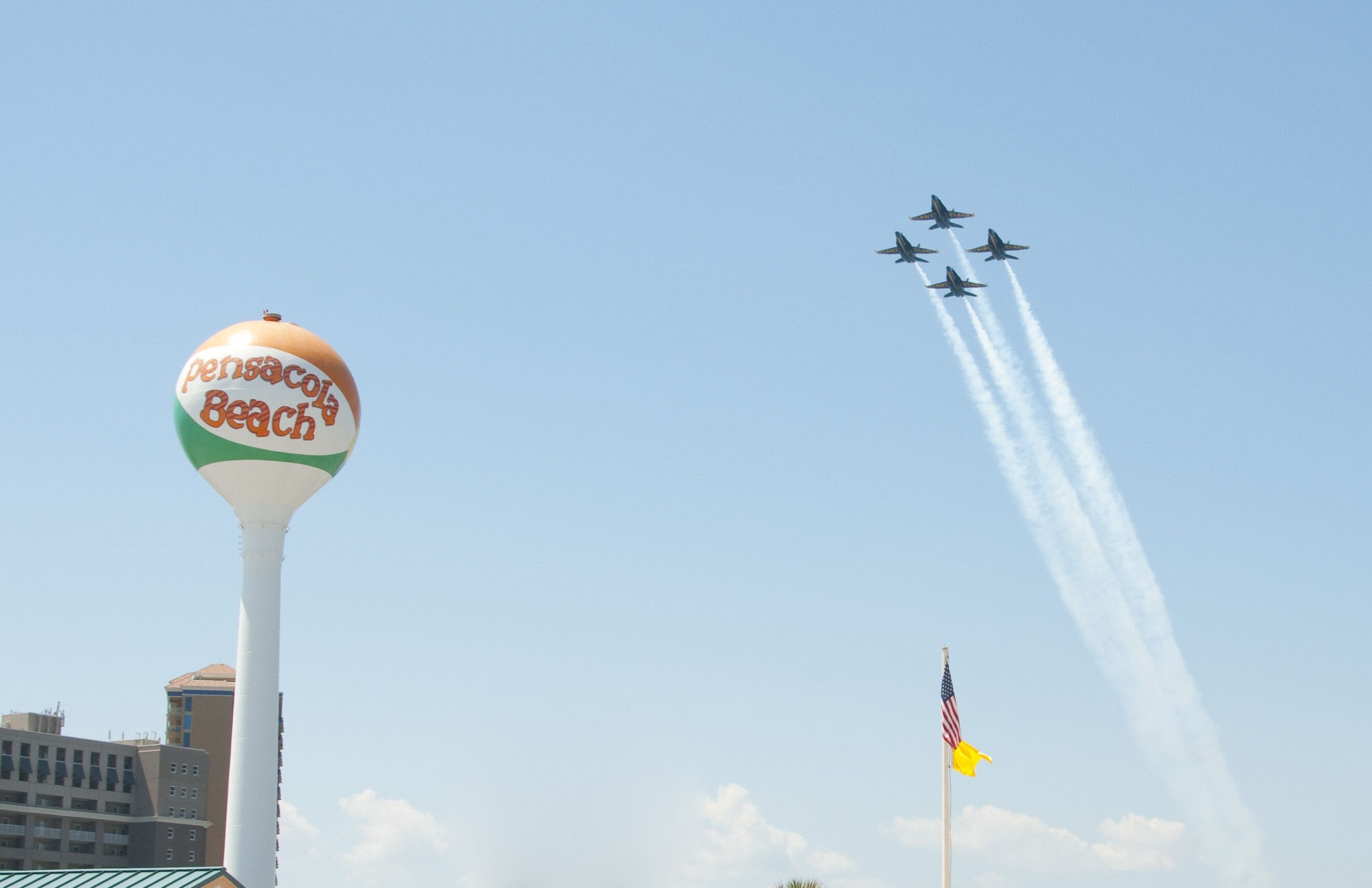 A water tower with "Pensacola Beach" written on it, a flagpole with an American flag, and four jets performing a flight formation.