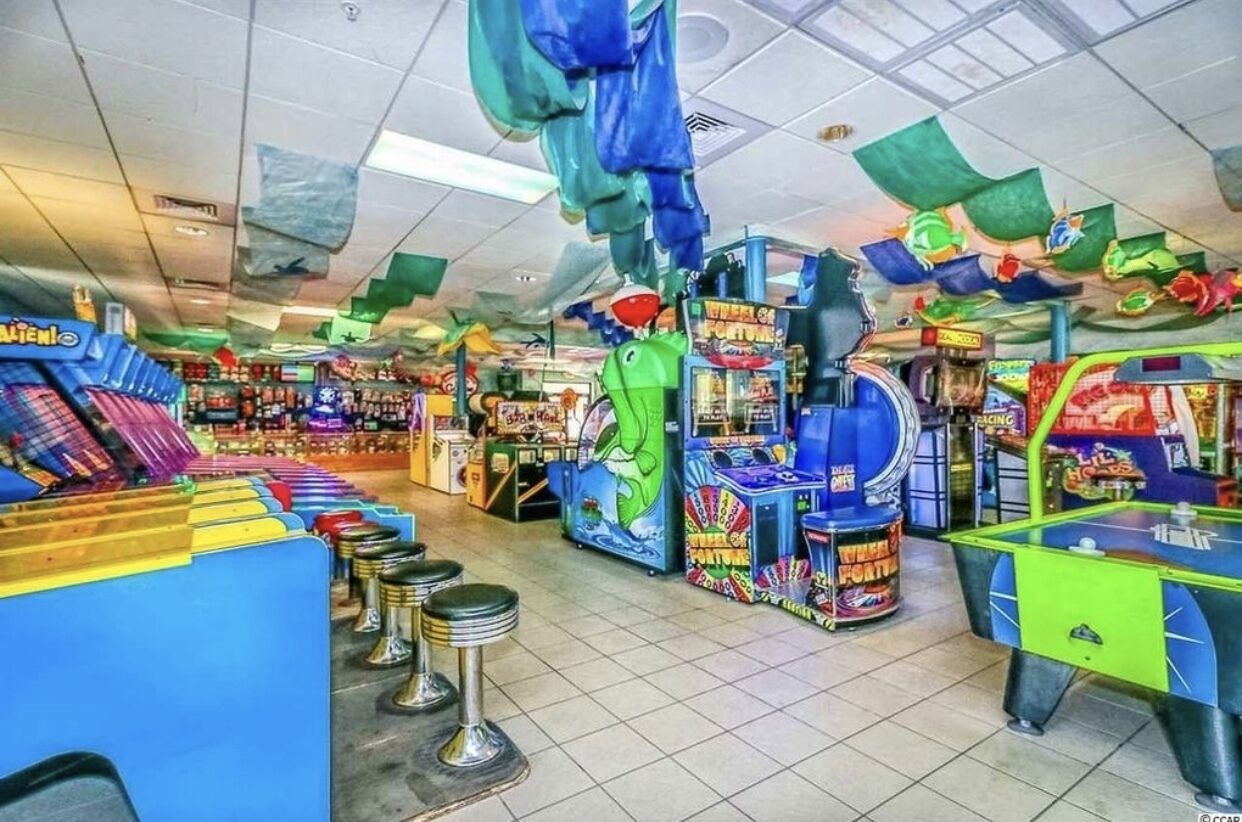 A brightly lit arcade with various gaming machines, including a green dinosaur-themed game and a blue racing game.