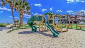 A beachside playground with a slide, surrounded by palm trees and a building with balconies.