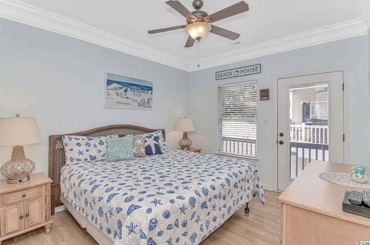 Ein Schlafzimmer mit Bett, Deckenventilator, Nachttisch, Lampe, Kommode und einem Fenster mit Jalousien.