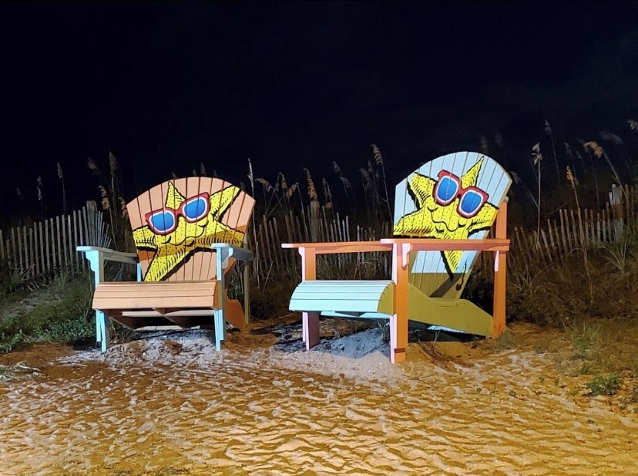 Two beach chairs with cartoon faces and sunglasses, placed on a sandy surface with a wooden fence in the background.