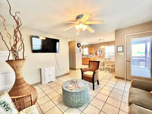 A living room with a TV, a ceiling fan, a dining area with chairs, a kitchen in the background, and a large window.