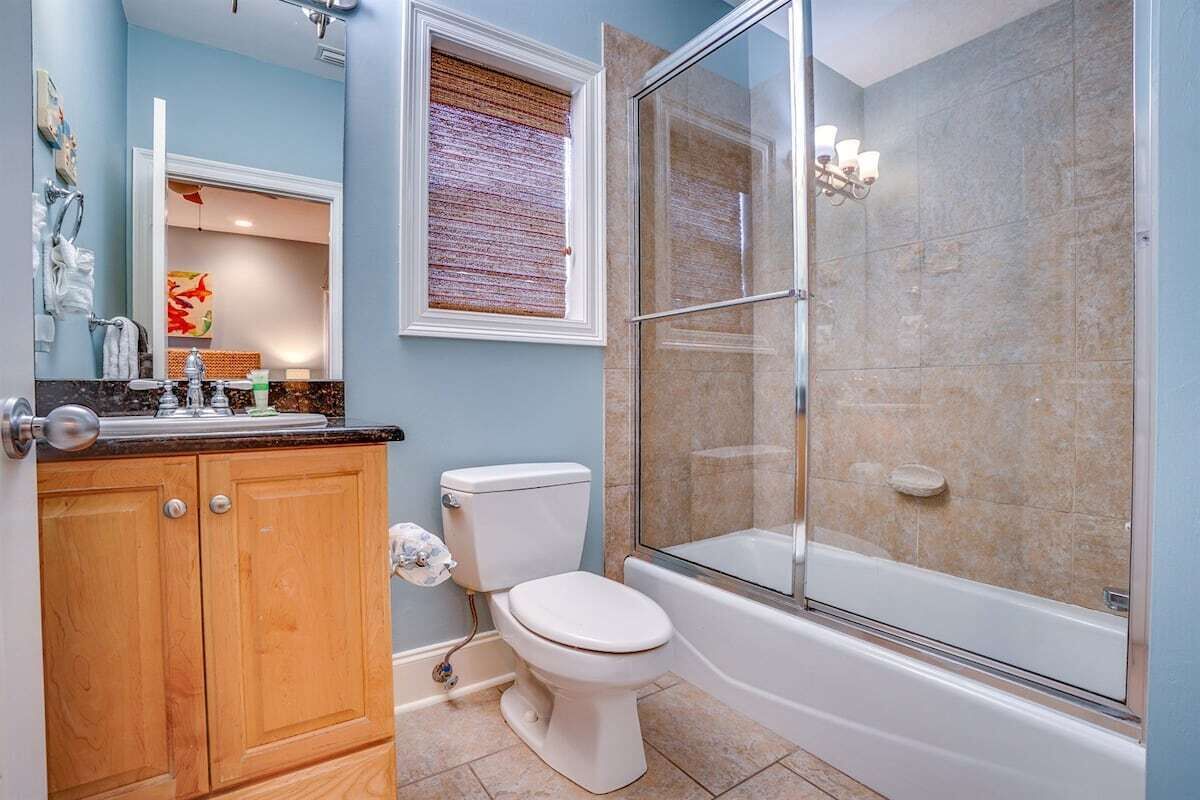 A bathroom with a toilet, a shower with a glass door, wooden cabinets, and a window with blinds.