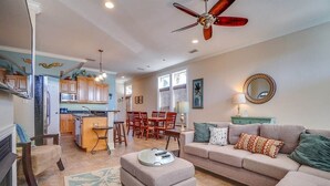 A spacious living room with a sofa, ottoman, and a ceiling fan. A kitchen with wooden cabinets and a dining area with chairs is visible in the background. - 🌈Amzng Gulf view, 4 Bd/3.5 Bath Home,🏖Pvt Heated Pool☀️,🌴Just Steps to Gulf🌴 (Panama City Beach)
