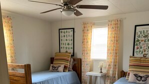 Una camera da letto con due letti in legno, una ventola a soffitto, tende floreali e un comodino con una lampada.