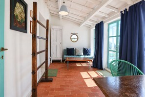Una habitación con puerta de madera, una silla verde, un sofá con cojines, un espejo redondo y una ventana con cortinas.