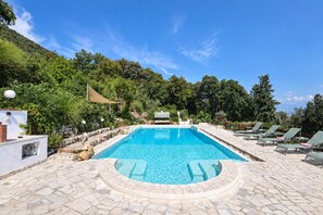 Une piscine rectangulaire entourée d’une terrasse en pierre, un espace de détente avec des fauteuils et une vue sur un paysage montagneux.