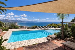 A swimming pool with a view of the sea and mountains, surrounded by lounge chairs and a shaded area.