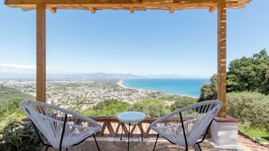 A wooden pergola with two white wicker chairs and a small table overlooking a coastal view.