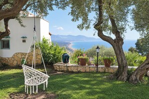 Un edificio blanco con base de piedra, una silla colgante, plantas en macetas y vistas al mar y a las montañas.