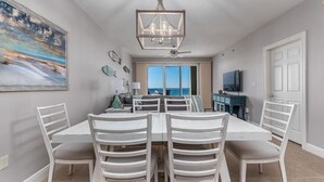 Un coin repas avec une table et des chaises blanches, un lustre, un ventilateur de plafond et un grand tableau accroché au mur.