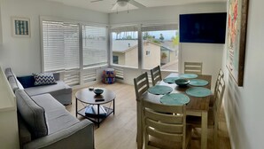 A living room with a sofa, a dining table with chairs, a television, and large windows with blinds. - San Clemente's "Jewel" - Blue Whale Inn #D (San Clemente)