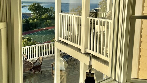 Une vue depuis une fenêtre ornée d’une balustrade blanche, donnant sur un court de tennis et une étendue d’eau.