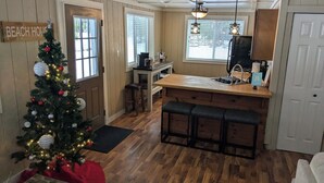 Une cuisine aux murs en bois, un îlot central également en bois avec des tabourets de bar, un sapin de Noël et une pancarte sur laquelle on peut lire : « MAISON DE PLAGE ».