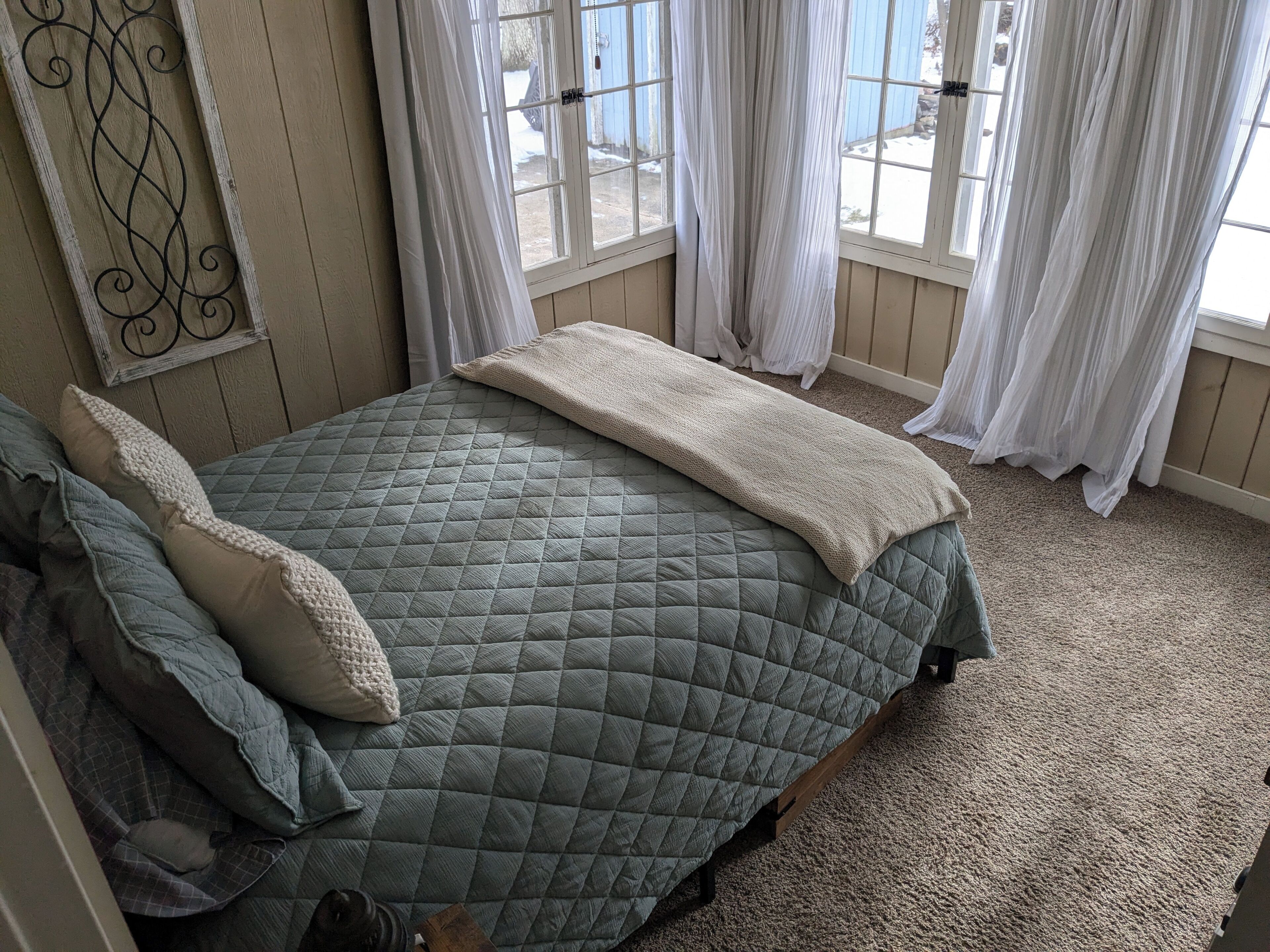 A bedroom with a bed, pillows, a quilted bedspread, and a view of the outdoors through a window with curtains.