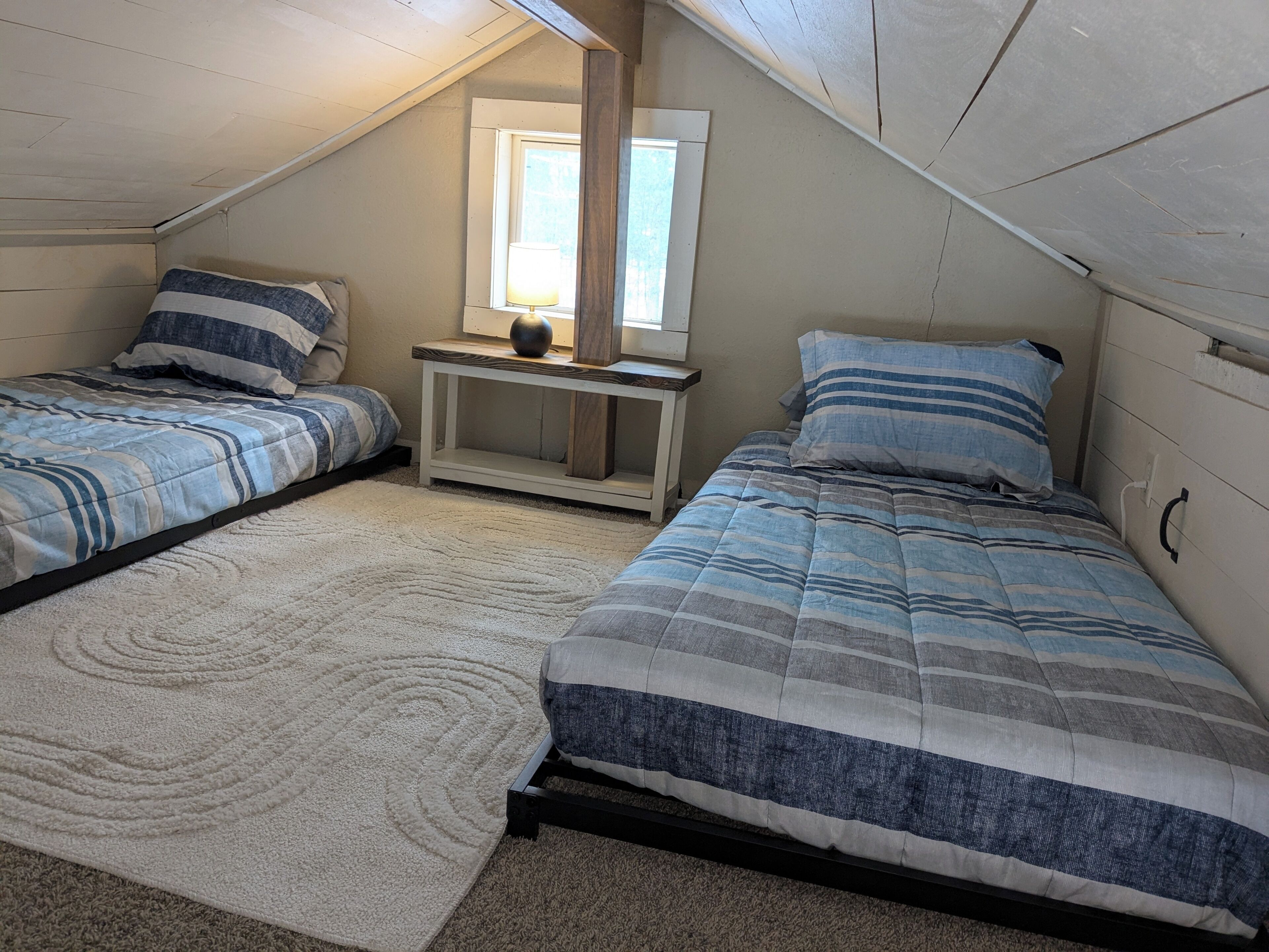 Ein Schlafzimmer mit zwei Betten, einem kleinen Nachttisch mit Lampe, einem Fenster und einem weißen Teppich.