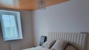 Un dormitorio con cama, mesita de noche, silla, ventana y lámpara en el techo.