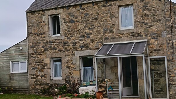 Una casa de piedra con techo inclinado, puerta de cristal y un pequeño jardín con plantas en macetas.