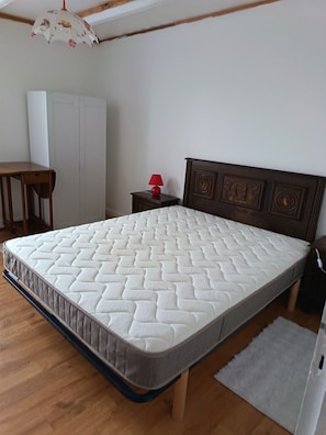 A bedroom with a wooden floor, a bed with a quilted mattress, a nightstand with a red lamp, a wooden headboard, and a wardrobe.