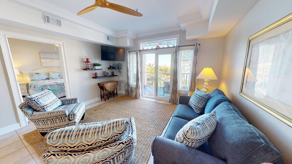 A spacious living room with a large sofa, patterned armchairs, a coffee table, and a TV. There is a balcony with a view, and the room has a modern design with white walls and ceiling.