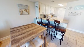 A kitchen with a wooden table and four chairs, white cabinets, a clock, and a wall with a nautical-themed artwork.