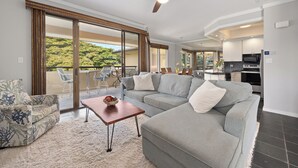 Una sala moderna con un sofá seccional gris, una pequeña mesa de centro de madera y un gran ventanal corredero que da a un balcón con mobiliario exterior.