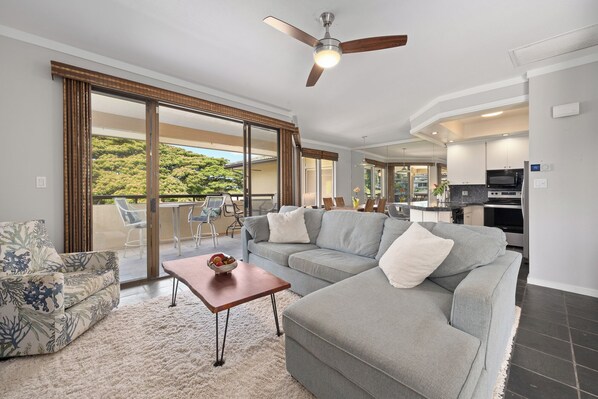 A modern living room with a grey sectional sofa, a small wooden coffee table, and a large sliding glass door leading to a balcony with outdoor furniture.