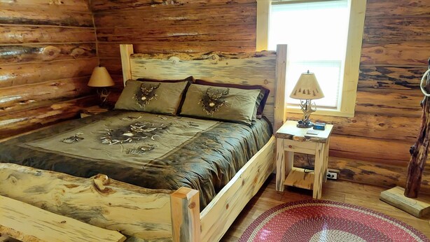 Un dormitorio de cabaña de madera con una cama, dos lámparas, una mesita y una ventana.