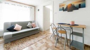 A modern living room with a grey sofa, a wooden dining table with metal chairs, and a large window with blinds.