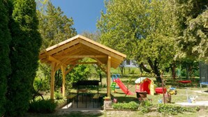 A wooden pavilion with a table and benches, surrounded by trees and a playground with slides.