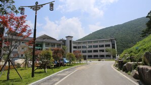 A road leading to a modern building with multiple floors, surrounded by greenery and hills.