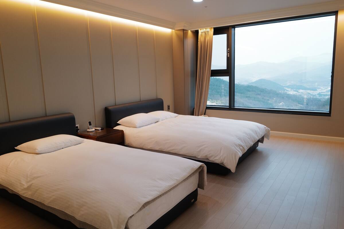 A hotel room with two beds, a nightstand, and a large window offering a mountain view.