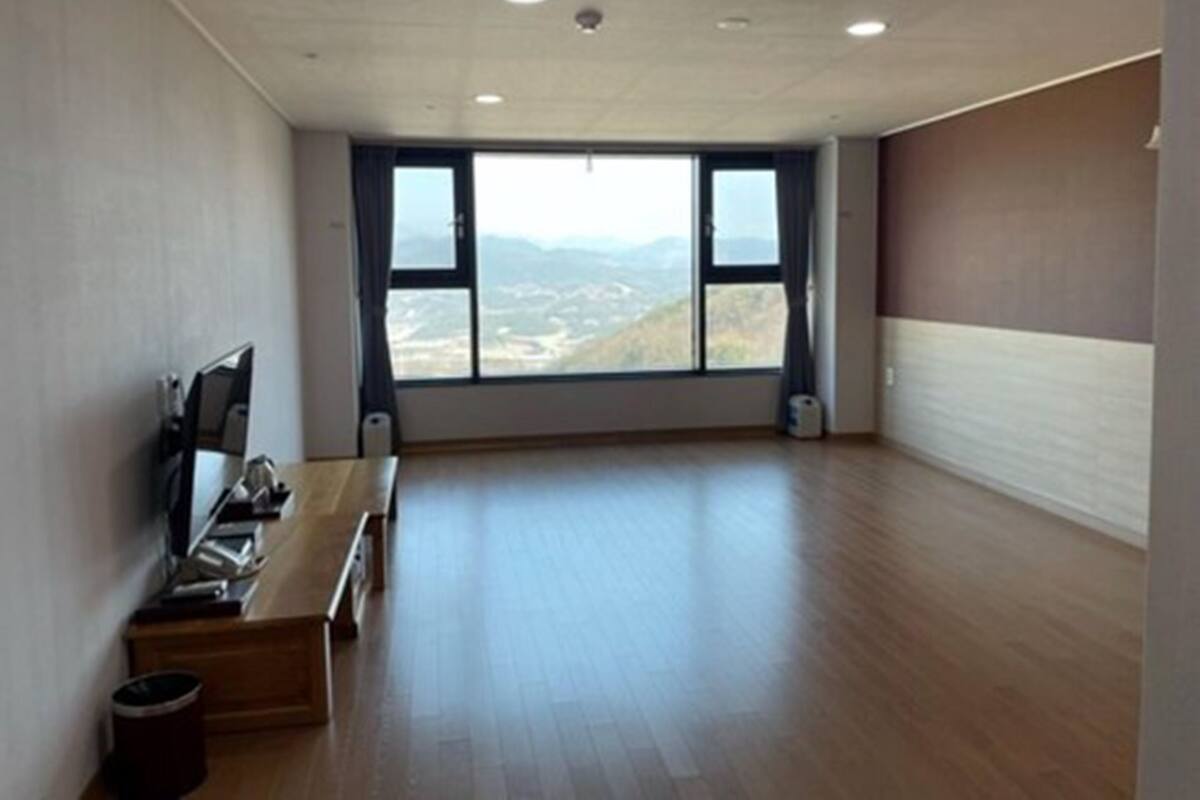 A spacious room with a large window offering a view of distant hills. A wooden bench and a small trash bin are visible.