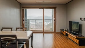 A modern living room with a wooden floor, a dining table with chairs, a TV on a wooden stand, and a sliding glass door leading to a balcony with a view of mountains.