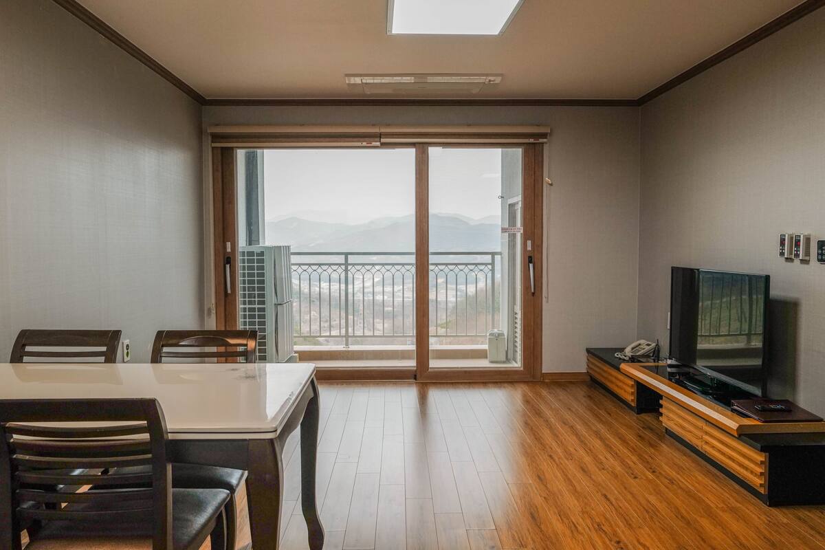 A modern living room with a wooden floor, a dining table with chairs, a TV on a wooden stand, and a sliding glass door leading to a balcony with a view of mountains.