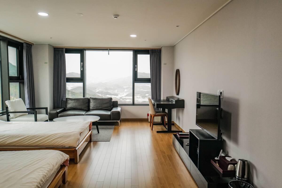 A modern hotel room with two beds, a sofa, a desk, and a large window offering a city view.