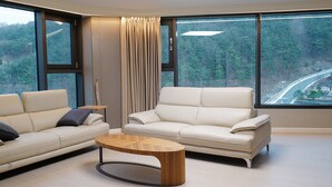 A modern living room with two sofas, a coffee table, and large windows offering a view of a forest and a road.