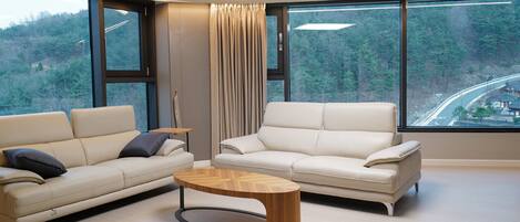 A modern living room with two sofas, a coffee table, and large windows offering a view of a forest and a road.