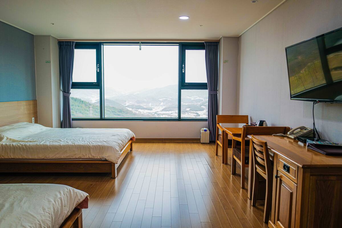 A hotel room with two beds, a desk, a chair, a TV, and a large window with a view of the mountains.