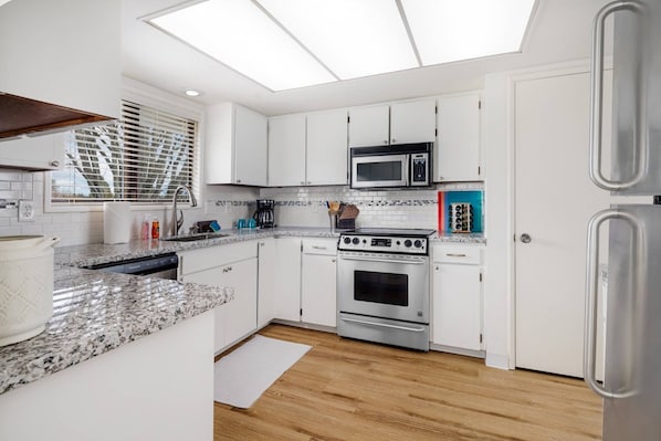 Une cuisine moderne avec des meubles blancs, une cuisinière en acier inoxydable, un four à micro-ondes et une grande fenêtre équipée de stores.