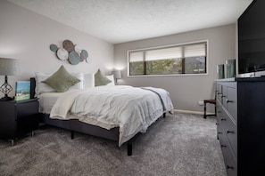 Une chambre à coucher comprenant un lit, des tables de chevet, une commode et une fenêtre avec des stores.