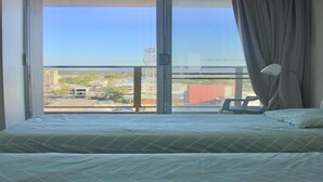 Een hotelkamer met een grote schuifpui naar een balkon, een bed met een bedspreide met groen patroon, en een witte stoel.